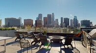 Located at 1100 West Temple Street in Los Angeles, this rooftop terrace offers an exceptional outdoor amenity space with sweeping, unobstructed views of the Downtown Los Angeles skyline. The terrace is thoughtfully divided into distinct zones, each designed for a different outdoor experience. A central fire pit seating area serves as the social heart of the space, surrounded by a curated arrangement of lounge chairs and wire accent seating, ideal for gathering under the open sky. Adjacent to the fire pit, a dining and lounge area features a sleek white table and a mix of contemporary seating options. An outdoor kitchen equipped with a built-in grill provides everything needed for alfresco cooking and entertaining. On the opposite end of the terrace, a lush artificial turf lawn offers a casual recreational zone complete with a ping-pong table, lounge chairs, a wall-mounted flat-screen television, and a shaded umbrella seating area — perfect for relaxed outdoor leisure. Mature trees, ornamental grasses, drought-tolerant shrubs, and vibrant red foliage plants are integrated throughout the terrace, adding color, texture, and a sense of privacy. The large-format tile paving, modern architectural detailing, and abundant Southern California sunshine create a polished yet inviting outdoor environment.