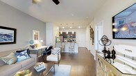 This thoughtfully designed single-floor residence showcases a seamless open-concept layout that effortlessly connects the living, dining, and kitchen areas. Rich hardwood-style flooring runs throughout, lending warmth and continuity to the entire space. The modern kitchen is a standout feature, equipped with stainless steel appliances, dark cabinetry, and a center island with bar seating illuminated by elegant pendant lighting — perfect for both casual dining and entertaining. Recessed lighting overhead ensures the space remains bright and welcoming at all hours. The living area is generously proportioned, offering ample room for comfortable furnishings, and is complemented by a ceiling fan that adds both function and character. Soft, neutral wall tones in muted greens and whites create a calm, contemporary backdrop that pairs well with the curated décor. A hallway extends toward additional rooms, offering a sense of depth and private separation from the main living space. Decorative wall accents and thoughtful styling details throughout reflect a refined aesthetic, making this residence a harmonious blend of modern functionality and inviting comfort.