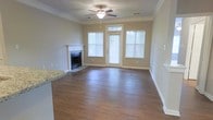This single-floor residence offers a welcoming and well-appointed living environment designed with both comfort and functionality in mind. The main living area is a standout feature, showcasing rich hardwood flooring that extends throughout the space, creating a warm and cohesive aesthetic. Crown molding lines the ceiling, adding a refined architectural detail that elevates the overall character of the room.

A classic fireplace with a clean white mantle serves as a natural focal point, providing both visual appeal and a cozy atmosphere. Adjacent to the fireplace, large windows fitted with white blinds invite generous amounts of natural light, brightening the interior while offering views of the outdoors. A glass-paneled door with a transom window above further enhances the flow of light and provides convenient access to the exterior.

The ceiling fan with integrated lighting ensures year-round comfort and practical illumination. A glimpse of granite countertop visible at the edge of the frame hints at a well-finished kitchen nearby, suggesting consistent quality finishes throughout the home. The neutral wall tones complement the warm flooring and trim, creating a versatile backdrop suitable for a variety of interior styles. Overall, the space presents a clean, move-in-ready environment with timeless design details.