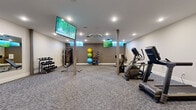 This fitness center offers a comprehensive and thoughtfully organized exercise space designed to support a wide range of workout routines. The room is finished with durable, patterned carpet flooring and bright recessed lighting complemented by a centrally mounted ceiling ventilation unit, creating a clean and comfortable environment for physical activity.

The cardio zone is well-stocked with treadmills, an elliptical trainer, and a stationary bike, all arranged to allow for easy circulation and use. A dedicated strength training area features a multi-function cable machine, a tiered dumbbell rack with a full range of free weights, a weight bench, and a collection of exercise and medicine balls neatly organized on a storage rack. Structural support columns are integrated throughout the space without impeding functionality.

Multiple large wall-mounted flat-screen TVs are positioned throughout the room, ensuring visibility from nearly any workout station. Full-length mirrors line one wall, enhancing the sense of openness and allowing users to monitor their form during exercise. The entry area features glass-panel doors that connect to the broader facility, contributing to an open and accessible feel. The overall design is modern and functional, providing users with a well-rounded fitness experience in an efficiently utilized space.