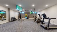 This fitness center offers a comprehensive and thoughtfully organized exercise space designed to support a wide range of workout routines. The room is finished with durable, patterned carpet flooring and bright recessed lighting complemented by a centrally mounted ceiling ventilation unit, creating a clean and comfortable environment for physical activity.

The cardio zone is well-stocked with treadmills, an elliptical trainer, and a stationary bike, all arranged to allow for easy circulation and use. A dedicated strength training area features a multi-function cable machine, a tiered dumbbell rack with a full range of free weights, a weight bench, and a collection of exercise and medicine balls neatly organized on a storage rack. Structural support columns are integrated throughout the space without impeding functionality.

Multiple large wall-mounted flat-screen TVs are positioned throughout the room, ensuring visibility from nearly any workout station. Full-length mirrors line one wall, enhancing the sense of openness and allowing users to monitor their form during exercise. The entry area features glass-panel doors that connect to the broader facility, contributing to an open and accessible feel. The overall design is modern and functional, providing users with a well-rounded fitness experience in an efficiently utilized space.