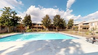 This community amenity area is centered around a generously sized rectangular swimming pool with clear, blue-tinted water, accessible via multiple entry points including handrail-equipped steps. The pool is surrounded by an expansive concrete sun deck that provides ample space for lounging and recreation. A black wrought-iron perimeter fence encloses the pool area, offering both safety and a defined boundary between the aquatic space and the surrounding grounds.

Mature shade trees are distributed throughout the property, adding natural greenery and a sense of established character to the outdoor environment. Picnic-style tables with attached seating are positioned along the deck, providing a communal gathering area for residents. The surrounding multi-story residential buildings feature earth-toned stucco exteriors, wood-accented balconies, and exterior staircases, contributing to a cohesive architectural aesthetic.

A dedicated mailroom corridor serves the community, featuring a skylight overhead that introduces natural light into the passage. Banks of individual mailboxes line one wall, and the corridor connects to building entry doors, keeping resident mail services centralized and convenient. The overall ambiance of the outdoor space is open and sun-drenched, suited for warm-weather enjoyment.