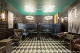 Located at 902 W Peachtree St NW in Atlanta's Midtown neighborhood, this single-floor lounge space delivers a bold, sophisticated aesthetic defined by dramatic design choices throughout. The interior is enveloped in floor-to-ceiling glossy black subway tile, creating a reflective, cave-like ambiance that sets a distinctly moody tone.

The flooring throughout is a striking black-and-white geometric patterned tile featuring large oval and diamond motifs, providing a graphic contrast to the dark walls. Seating is arranged across several distinct zones: plush leather club chairs are grouped in conversational clusters, while velvet-upholstered banquette booths flank round pedestal tables in a more intimate corner, lit by globe pendant fixtures overhead.

A built-in bar area along one wall features a compact beverage refrigerator and shelved glassware, accompanied by a wall-mounted flat-screen television and flanked by brass sconce lighting. Large-format black-and-white photographic artworks — depicting crystal decanters and suited figures — are displayed prominently throughout, reinforcing the space's refined, speakeasy-inspired character. Teal-painted ceilings and gold drapery add further depth and warmth to the overall atmosphere.