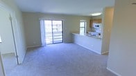 This single-floor unit is designed with an open-concept layout that seamlessly connects the living area and kitchen. The spacious living room is finished with plush wall-to-wall carpeting in a neutral tone, providing a comfortable and clean foundation throughout the space. Soft beige walls contribute to a bright, welcoming ambiance that pairs well with the abundant natural light streaming in from multiple windows.

A large sliding glass door dominates one wall of the living area, offering direct access to an outdoor balcony and framing views of the surrounding exterior. This feature enhances the sense of openness and keeps the interior feeling airy and well-lit throughout the day.

The kitchen is positioned adjacent to the living space, separated by a raised peninsula counter with a light-toned surface that doubles as a casual breakfast bar or serving area. Warm wood-toned cabinetry lines the kitchen walls, providing ample storage, while a white microwave and range are integrated neatly into the layout. A dedicated window above the kitchen brings in additional natural light, keeping the cooking area bright and functional.

Overall, the unit presents a clean, practical, and well-lit living environment suitable for everyday comfort.