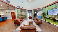Located at 2735 N Monroe St in Tallahassee, Florida, this single-floor community clubhouse offers a generously sized, open-plan interior designed to accommodate a variety of social and recreational activities. The space is defined by soaring high ceilings and an abundance of natural light streaming through large windows and multiple sets of French doors that open to outdoor decking areas, providing views of the surrounding landscape.

A striking design feature throughout is the use of large-scale, floor-to-ceiling nature-inspired wall murals depicting lush forest scenes, which lend a distinctive and immersive ambiance to each zone. The main lounge area is anchored by comfortable seating arrangements, including leather sofas and accent chairs, centered around coffee tables atop wood-look flooring that runs consistently throughout the space.

A dedicated dining and café area features bistro-height tables paired with colorful seating and elegant tear-drop pendant lighting in aqua glass. A grand piano serves as a standout focal point in one section, complemented by a wall-mounted flat-screen television. Additional areas include a workspace or business center with desks and shelving, as well as a central service counter. The curved architectural detailing in the entry zone adds a refined, welcoming character to the overall design.