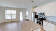 This single-floor space showcases a clean, modern design centered around an open-concept layout that seamlessly connects the kitchen and living area. The kitchen is a standout feature, highlighted by a large central island with a sleek quartz-style countertop and an undermount sink with a pull-down faucet. White shaker-style cabinetry lines the walls, providing ample storage, while stainless steel appliances — including a French door refrigerator, electric range, and built-in microwave — complete the contemporary aesthetic. Recessed lighting throughout the space ensures consistent, even illumination. Light wood-look flooring runs across the entire floor, contributing to a warm yet modern atmosphere. The living area beyond the kitchen benefits from multiple windows that allow generous natural light to fill the space, enhancing the open and airy feel. A glass-panel door leads to the exterior, adding to the brightness and providing outdoor access. The overall color palette — whites, soft neutrals, and light wood tones — creates a cohesive, inviting environment suited to modern living.
