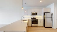 Located at 123 Water St in Norwalk, Connecticut, this contemporary single-floor residence showcases a clean, modern design throughout. The open-concept kitchen is a standout feature, outfitted with crisp white cabinetry, a spacious quartz countertop peninsula, and a full suite of stainless steel appliances including a gas range, built-in microwave, dishwasher, and refrigerator. Stylish pendant lighting hangs above the peninsula, adding warmth to the bright, airy space. Light-toned hardwood flooring flows through the main living areas, enhancing the sense of openness and continuity. Natural light pours in through large windows, creating a welcoming ambiance throughout the day. The bathroom is elegantly finished with marble-style tile surrounding the soaking tub and shower combination, complemented by neutral floor tile for a cohesive, refined look. The bedroom features plush carpeting underfoot and floor-to-ceiling sliding glass doors that open onto a private balcony, offering expansive views of the surrounding waterfront and cityscape. The overall design aesthetic is fresh and minimalist, with white walls and thoughtful finishes that create a polished, move-in-ready living environment.