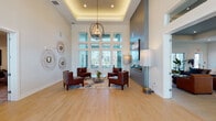 This thoughtfully designed single-floor amenity space combines elegant architecture with practical functionality across a generous, open layout. The centerpiece is a dramatic lounge area featuring soaring ceilings with an illuminated tray ceiling detail, a striking orb chandelier, a wall-mounted fireplace framed in dark tile, and a large window wall that floods the room with natural light. Rich hardwood-style flooring runs throughout, complementing the warm, neutral palette.

A bright, open social area extends toward floor-to-ceiling sliding glass doors that open directly to an outdoor terrace, where palm trees and open skies create a seamless indoor-outdoor connection. A sculptural art piece and lush tropical plants add character to this inviting gathering space.

The dining room offers an intimate setting with a round table, pendant lighting, built-in cabinetry, and curated wall art. Adjacent is a fully equipped kitchen featuring white shaker cabinetry, open wood shelving, subway tile backsplash, stainless steel appliances, and a large island with bar seating.

A wide, light-filled corridor connects private office and conference spaces, each appointed with modern furnishings and patterned area rugs. A cozy lounge nook with a plush sectional and abstract artwork provides a relaxed retreat. The welcoming entry foyer features a tray ceiling, decorative console, and elegant pendant fixture, setting a polished tone throughout.