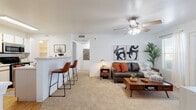 This single-floor residence offers a thoughtfully designed layout that balances comfort and functionality throughout. The open-concept living area is carpeted and bathed in natural light from large windows and a glass door, which provide outdoor views and a bright, airy atmosphere. A ceiling fan with integrated lighting keeps the space comfortable year-round. The living room flows seamlessly from a kitchen peninsula fitted with a breakfast bar and leather-upholstered counter stools, creating an ideal space for casual dining and entertaining.

The kitchen features light wood cabinetry, stainless steel appliances including a range and over-the-range microwave, a double-basin sink, and light-toned countertops. Wood-look flooring runs throughout the kitchen, and a walk-in pantry provides ample storage. A hallway connects the main living spaces to the private areas of the home.

The bedroom is generously sized with plush carpeting, a window that invites natural light, and enough room to comfortably accommodate a large bed and additional furnishings. The bathroom includes a vanity with under-sink cabinetry, a well-lit mirror, and wood-look flooring, offering a clean and practical design. Overall, the space presents a cohesive, modern aesthetic with warm tones throughout.