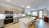 This single-floor residence offers a well-designed open-concept layout connecting the kitchen and living areas. The kitchen is outfitted with flat-front cabinetry in a warm gray-taupe finish, white quartz countertops, and a full suite of stainless steel appliances including a refrigerator, range, microwave, and dishwasher. A large central island with seating provides additional prep space and casual dining. Pendant lighting above the island adds a modern touch, complementing under-cabinet lighting along the perimeter counters. A textured herringbone tile backsplash adds visual interest.

The living area extends naturally from the kitchen and is bathed in natural light from large windows and a glass door that opens to a balcony with urban views. Light wood-look flooring runs throughout the main living spaces, contributing to a cohesive, airy feel. A ceiling fan keeps the space comfortable.

The bedroom is generously sized, featuring a window with natural light, ceiling fan, and neutral walls. The bathroom connects directly to the bedroom and includes a wood-toned vanity with white quartz countertop, a lighted mirror, and a tub-shower combination with white tile surround.
