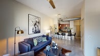 This one-bedroom, one-bathroom residence in Houston, TX offers a thoughtfully designed open-concept floor plan that blends industrial and modern aesthetics. Polished concrete floors flow throughout the main living areas, complementing the exposed ductwork that adds an urban loft character to the space. The living room is bright and welcoming, featuring soaring ceilings, a ceiling fan, and large sliding glass doors that open onto a private balcony with views of lush surrounding greenery. The kitchen is well-appointed with rich wood cabinetry, granite countertops, a tile backsplash, and black appliances including a refrigerator and dishwasher. A curved granite breakfast bar with bar seating connects the kitchen to the living area, creating a seamless entertaining space. Built-in open shelving provides additional storage and display opportunities. The bedroom is a comfortable retreat with a large window that invites natural light and views of the greenery outside. The full bathroom features a tiled soaking tub and shower combination with classic black accent tile detailing, polished concrete flooring, and access to a walk-in closet with built-in shelving. Track lighting throughout adds functional illumination to this well-designed Houston residence.