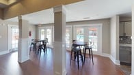 This single-floor amenity space is thoughtfully designed for social interaction and recreation, combining distinct functional zones within an open, well-appointed layout. The centerpiece of the space is a generously sized billiards room, anchored by a full-size pool table with rich wood detailing and a black felt surface. Above, a coffered ceiling with decorative beams adds architectural depth, while a statement pendant light fixture illuminates the table. Wall-mounted cue racks and a flat-screen TV enhance the room's game-room functionality. Adjacent to the billiards area, an interior pass-through window connects to a bar and lounge area, where bar-height seating and decorative artwork create a casual, social atmosphere with pendant lighting overhead.

The lounge and dining area features high-top tables and metal bar stools arranged around white decorative columns, with rich hardwood flooring running throughout the space. Large French doors and oversized windows line the exterior wall, flooding the interior with natural light and offering direct views of an outdoor pool and surrounding patio. The textured ceiling, neutral taupe walls, and white trim millwork provide a clean, cohesive aesthetic throughout, making this space ideal for community gatherings and leisure activities.