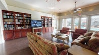 This inviting single-floor space is designed for comfort and community, blending a library and lounge atmosphere seamlessly. The focal point of the room is an impressive wall of rich cherry wood built-in bookshelves, fully stocked with an extensive collection of books, flanking a large flat-screen television — perfect for both quiet reading and group entertainment. Ceiling fans are thoughtfully positioned throughout, contributing to a well-ventilated and comfortable environment. The living area is furnished with generously sized, patterned sofas and armchairs in warm earth tones, arranged around an ornately carved coffee table, creating an inviting space for conversation and relaxation. Dark hardwood flooring runs throughout, adding a sense of warmth and elegance to the space. Large windows along one wall allow abundant natural light to pour in, offering views of the outdoor surroundings and creating a bright, airy ambiance. The overall design strikes a balance between functional and welcoming, making this a comfortable gathering space well-suited for reading, socializing, or simply unwinding.
