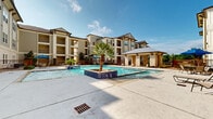 Located at 31623 Waller Tomball Rd in Waller, Texas, this outdoor amenity area is the centerpiece of a well-appointed residential community. The space is anchored by a generously sized swimming pool with clear blue water, complemented by an attached spa featuring decorative tiled surrounds and a palm tree centerpiece that adds a resort-like character to the environment.

The pool deck is finished in light-toned concrete, offering ample room for lounge chairs and outdoor furniture, providing residents with comfortable spaces to relax and enjoy the Texas sunshine. A standout feature is the covered cabana, constructed with natural stone columns and a warm wood-paneled ceiling fitted with ceiling fans, offering a shaded retreat directly adjacent to the pool. The tile flooring within the cabana extends the resort aesthetic and creates a refined outdoor gathering area.

The surrounding multi-story buildings feature a mix of stucco siding, stone accents, pitched rooflines, and private balconies, creating an attractive architectural backdrop. Wrought iron fencing encloses the pool area, enhancing both security and visual appeal. Landscaping throughout includes palm trees, ornamental plantings, and decorative rock beds that frame the space cohesively. The overall ambiance is one of a well-maintained, community-focused outdoor retreat designed for relaxation and leisure.