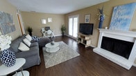 This two-story home offers a thoughtfully designed layout that balances comfort and functionality. The main living area immediately draws attention with its rich, dark hardwood flooring that extends throughout the space, creating a warm and cohesive aesthetic. Neutral, warm-toned walls provide a versatile backdrop that complements a variety of décor styles.

The living room serves as the heart of the home, highlighted by a classic white-framed fireplace that adds both architectural character and a cozy focal point. Large windows and glass-paned doors allow generous natural light to pour into the room, creating a bright and airy atmosphere. The space is well-proportioned, comfortably accommodating a full seating arrangement alongside a dedicated workspace or desk area, demonstrating its flexible use of space.

The open layout promotes a natural flow between living and functional zones, making it ideal for both everyday living and entertaining. Tasteful decorative touches, including indoor greenery and carefully placed artwork, contribute to an inviting ambiance throughout. The combination of the fireplace, hardwood floors, and abundant natural light gives the home a timeless, welcoming character. Spanning two floors, this property offers ample room to accommodate a variety of lifestyle needs with a classic, well-maintained design.