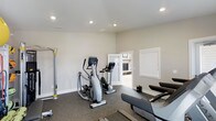 This single-floor fitness space is thoughtfully designed to support a well-rounded exercise routine. The room features durable rubber gym flooring that provides traction and cushioning underfoot, making it well-suited for a variety of workout activities. Recessed ceiling lighting ensures the space is evenly and brightly illuminated throughout.

The room is equipped with multiple cardio machines, including an elliptical trainer, a stationary exercise bike, and a rowing machine, offering users a range of low-impact cardiovascular workout options. A vertical workout station along one wall adds further versatility for strength and stretching exercises.

The walls are finished in a clean, neutral tone that contributes to a focused and calm workout environment. A window along one wall allows for natural light to filter in, enhancing the overall ambiance of the space. An open doorway connects the fitness room to an adjacent lounge or common area, providing convenient access and promoting a seamless flow between spaces.

The overall design is clean, functional, and modern, making this fitness room a practical and well-appointed space for daily exercise and physical wellness.