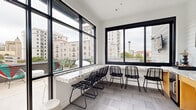 This contemporary rooftop space seamlessly blends an interior kitchen area with a sprawling outdoor terrace, offering a sophisticated urban retreat with striking city views. The interior kitchen features large, black-framed floor-to-ceiling windows that flood the space with natural light while framing impressive views of the surrounding cityscape, including a recognizable tower landmark on the horizon. A continuous marble-topped counter runs along the windowed walls, complemented by sleek black wire-frame chairs, creating a functional yet stylish bar-style seating arrangement. Light-toned tile flooring ties the interior together with a clean, modern aesthetic. The expansive outdoor terrace extends the usable space considerably, designed for both relaxation and entertainment. It is equipped with a wall-mounted flat-screen television, built-in lounge seating, a large white dining table with wire chairs, and colorful woven accent chairs that add a vibrant touch. Lush potted cacti and greenery are thoughtfully arranged throughout the terrace, adding a natural contrast to the urban surroundings. Perimeter railings ensure open, unobstructed panoramic views of the city skyline in multiple directions, making this rooftop an ideal setting for gatherings or quiet enjoyment above the city.