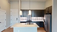 This well-appointed one-bedroom, one-bathroom unit in Austin, TX offers a thoughtfully designed single-level layout spanning just over 1,000 square feet. The open-concept living and kitchen area is anchored by large steel-framed glass doors that flood the space with natural light and provide direct outdoor access. Wood-look flooring runs throughout the main living areas, complemented by neutral gray walls and a ceiling fan overhead.

The kitchen is a standout feature, showcasing dark slate-toned shaker cabinets that extend to the ceiling for ample storage, a white quartz countertop island with an undermount sink and matte black faucet, a tiled subway backsplash, stainless steel appliances including a refrigerator and range, and pendant lighting above the island.

The bedroom offers carpeted flooring, a large window with outdoor views, and generous wall space. The bathroom is finished with light tile flooring, a dark-framed vanity mirror, white quartz countertop, matte black fixtures, and a glass-enclosed walk-in shower with vertical subway tile. Recessed lighting throughout the unit adds to its clean, modern aesthetic, making it a functional and attractive space in the heart of East Austin.
