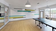 Located at 4050 Rocky Cir in Tampa, FL, this single-floor recreational facility is designed to deliver a dynamic, energetic environment for fitness and leisure. The space is defined by light hardwood flooring that runs throughout, white walls, and bold graphic wall art featuring horizontal stripes in teal, chartreuse, and black — giving the interior a modern, playful aesthetic.

The main open area serves as a multi-purpose room, highlighted by a full-size ping-pong table positioned beneath a series of large pendant lights with white dome shades. Natural light filters in through full-height windows and glass doors, keeping the space bright and inviting. The open layout allows for flexible use as both a game room and an activity space.

A dedicated fitness zone is equipped with a multi-tiered weight rack holding dumbbells, stability balls, kettlebells, and step platforms. A wall-mounted industrial fan provides ventilation, and a large floor-to-ceiling mirror panel visually expands the space while supporting exercise and movement activities. A flat-screen display is mounted directly to the mirror, offering functionality for guided workouts or entertainment.

The clean, contemporary design, open floor plan, and well-curated amenities make this space a comprehensive recreational hub suited for group fitness, casual games, and active recreation.