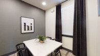 This conference room is a compact, well-appointed meeting space designed for focused collaboration. The room features a clean white rectangular table surrounded by sleek black chairs, providing seating for up to four people. The walls are finished in a two-tone palette, combining warm white with a bold charcoal accent wall, which adds depth and a contemporary aesthetic to the space. A framed typographic "hi" print adorns the accent wall, lending a welcoming and modern touch to the interior.

Natural light enters through a window dressed with dark blackout curtains and white horizontal blinds, allowing for flexible light control depending on the needs of the meeting. A wall-mounted flat-screen television is positioned for easy viewing, supporting presentations and collaborative work. Patterned carpet tiles cover the floor, adding visual texture while maintaining a professional atmosphere.

The room is enclosed by a full-length glass door clearly labeled "Conference Room," ensuring visibility from the adjacent common area while maintaining acoustic separation. Through the glass, a larger lounge area with additional seating and a pool table is visible, suggesting this conference room is part of a broader amenity floor. A small potted fern on the table adds a natural, inviting element to the otherwise clean and minimalist design.