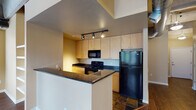 This single-floor loft-style unit showcases a compelling blend of industrial and contemporary design. Exposed concrete ceilings with visible steel beams and large metallic ductwork run throughout the space, lending it a distinct urban character. Rich, dark hardwood flooring flows seamlessly across the main living areas, providing a warm contrast to the industrial overhead elements.

The open-concept kitchen features light maple cabinetry, dark granite countertops, and black appliances including a refrigerator, gas range, and built-in microwave. Track lighting illuminates the kitchen efficiently, while tile flooring defines the cooking zone. A peninsula counter creates a natural divide between the kitchen and living area.

The spacious living area is bathed in natural light from multiple large windows and a glass-panel door that opens to an outdoor space, offering views of the surrounding urban environment. Built-in shelving adds both function and visual interest to one of the living room walls.

A hallway with pendant lighting connects the main living space to the private areas, where a full bathroom features a combined tub and shower, a vanity with maple cabinetry, and hardwood flooring. Additional closet storage is also accessible from the hallway. The overall ambiance is open, bright, and distinctly loft-inspired.