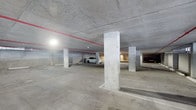 This single-level covered parking structure is built with a robust concrete construction, featuring a smooth poured-concrete floor, exposed concrete ceiling, and evenly spaced structural columns that define the layout of the space. The facility is designed to accommodate multiple vehicles, with clearly delineated parking bays marked directly on the floor surface. Overhead artificial lighting is distributed throughout the structure, ensuring consistent visibility across the entire floor. A prominent red fire suppression pipe runs along the ceiling, indicating a fully integrated safety system in compliance with standard building codes. The open-plan layout allows for ease of vehicle maneuverability, with wide driving lanes between rows of parking spaces. The neutral grey tones of the concrete surfaces throughout — from the floor to the ceiling and columns — give the structure a clean, utilitarian aesthetic. The overall design prioritizes functionality and safety, making it a practical and well-organized parking facility suited for residential or commercial use.