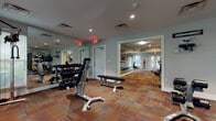 This fitness center is organized across a single floor and divided into distinct workout zones, creating a functional and spacious environment for a comprehensive exercise experience. The layout includes a dedicated cardio area lined with True-brand treadmills and stationary bikes, positioned along mirrored walls that visually expand the space and allow users to monitor their form. Large glass double doors open directly to an outdoor area, flooding the cardio zone with natural light and providing views of surrounding greenery.

A separate strength training area houses a full dumbbell rack, adjustable weight benches, and a cable machine, complemented by floor-to-ceiling mirrors along multiple walls. Recessed lighting throughout keeps the space well-lit, while the multi-toned, patterned carpet tiles add warmth and define each zone. Additional equipment, including a rowing machine and specialty strength machines, is distributed throughout, offering a well-rounded selection for varied fitness routines. Windows along the perimeter of the strength area bring in further natural light and outdoor views of lush foliage. The neutral gray walls and clean white trim contribute to a tidy, professional atmosphere throughout the facility.
