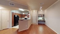 This single-floor residence offers a thoughtfully designed open-concept layout that seamlessly connects the kitchen and living areas. The kitchen is equipped with rich wood cabinetry, dark granite countertops, a black appliance suite including a refrigerator and range, and a breakfast bar peninsula that serves as both a prep space and casual dining area. Pendant lighting above the peninsula adds a warm, inviting glow, complementing the flush-mount and track lighting fixtures found throughout the space. Warm hardwood flooring runs continuously through the main living area, adding richness and visual continuity to the interior. Crown molding along the ceiling lends a polished, finished look to the space. A standout feature is the built-in corner desk nook, complete with open shelving above — a practical and charming detail that provides a dedicated workspace without sacrificing style. The entryway is clean and welcoming, anchored by a classic six-panel white door. Overall, the space strikes a balance between functionality and comfort, with well-planned built-ins, consistent finishes, and a layout that maximizes usability across every area of the floor plan.