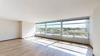 This single-floor unit showcases a contemporary design defined by clean lines, neutral tones, and an abundance of natural light. The living areas are anchored by expansive floor-to-ceiling windows that span entire walls, framing elevated panoramic views of the surrounding urban landscape and tree canopy. Light streams through the large glass panels, casting geometric patterns across the warm hardwood-style flooring that runs throughout the main living spaces.

The open layout provides a generous, flexible living area with smooth white ceilings and understated wall sconce lighting. The kitchen features a galley-style configuration equipped with rich, dark-stained wood cabinetry, dark granite countertops with a double stainless steel sink, stainless steel appliances including a refrigerator, gas range, and dishwasher, complemented by recessed lighting above.

The bathroom is generously proportioned, finished with neutral large-format tile flooring, a combination tub and shower with a handheld showerhead, grab bars, a toilet, and a dark wood vanity — reflecting an accessible and functional design. A dedicated walk-in closet offers wire shelving for organized storage. The overall ambiance is bright, airy, and modern, with a calm, minimalist aesthetic throughout.