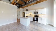 Located at 251 N Rose St in Kalamazoo, MI, this 1-bedroom, 1-bathroom unit showcases a compelling blend of industrial character and modern finishes. The open-concept living, dining, and kitchen area serves as the heart of the space, highlighted by soaring exposed wood beam ceilings with visible joists and ductwork — a nod to the building's architectural heritage. Light luxury vinyl plank flooring runs throughout, complementing the bright, neutral palette.

The kitchen is well-appointed with white shaker cabinetry, granite countertops, a subway tile backsplash, stainless steel appliances, and a central island with pendant lighting overhead. Natural light filters in through large windows and a glass-paned exterior door leading to a private balcony area.

A hallway connects the main living space to the private bedroom, which features exposed wood beams, large windows, and two generous closets with built-in hanging rods. The bathroom is finished with a walk-in tile shower with textured grey tile and matte black hardware, granite-topped vanity, and wire shelving for storage. An in-unit laundry area adds everyday convenience. This unit offers a distinctive urban-loft aesthetic with thoughtfully modern appointments throughout.