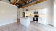Located at 251 N Rose St in Kalamazoo, MI, this 1-bedroom, 1-bathroom unit showcases a compelling blend of industrial character and modern finishes. The open-concept living, dining, and kitchen area serves as the heart of the space, highlighted by soaring exposed wood beam ceilings with visible joists and ductwork — a nod to the building's architectural heritage. Light luxury vinyl plank flooring runs throughout, complementing the bright, neutral palette.

The kitchen is well-appointed with white shaker cabinetry, granite countertops, a subway tile backsplash, stainless steel appliances, and a central island with pendant lighting overhead. Natural light filters in through large windows and a glass-paned exterior door leading to a private balcony area.

A hallway connects the main living space to the private bedroom, which features exposed wood beams, large windows, and two generous closets with built-in hanging rods. The bathroom is finished with a walk-in tile shower with textured grey tile and matte black hardware, granite-topped vanity, and wire shelving for storage. An in-unit laundry area adds everyday convenience. This unit offers a distinctive urban-loft aesthetic with thoughtfully modern appointments throughout.