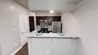 This single-floor residence offers a thoughtfully designed open layout that maximizes functionality and comfort. The kitchen serves as a central highlight, featuring rich dark-brown cabinetry that provides ample storage and creates a striking contrast against the neutral, light-toned walls. Stainless steel appliances — including a refrigerator, range, and built-in microwave — lend a sleek, contemporary feel to the space. The kitchen flows seamlessly into the living area, anchored by a peninsula with a granite-style countertop that doubles as a breakfast bar, complete with white bracket supports and seating for two. The countertop's light, speckled finish adds texture and elegance to the overall aesthetic. Dark hardwood-style flooring runs throughout, grounding the space with warmth and visual depth. A bifold-door closet is conveniently positioned near the kitchen, offering additional storage. The clean lines, neutral color palette, and cohesive design choices throughout give the space a modern, move-in-ready ambiance that balances practicality with style.