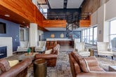 This upscale communal space combines industrial-modern design with warm, inviting elements across an open, single-floor layout. The main lounge area commands attention with its dramatic double-height ceilings, partially clad in rich warm wood paneling, and an abundance of natural light streaming through expansive floor-to-ceiling windows. These windows frame sweeping city skyline views, including prominent high-rise buildings, creating a seamless connection between the interior and the urban landscape beyond. A terrace with outdoor seating is visible through the glass, extending the living space outdoors.

The lounge is furnished with multiple generously sized leather sofas and armchairs arranged on a decorative area rug, providing ample seating for gathering and relaxation. A wall-mounted television is tucked beneath the wood ceiling feature, alongside pendant and track lighting that adds an industrial character. A sculptural black metal spiral staircase serves as a striking architectural focal point, providing access to a rooftop level above.

Adjacent to the lounge, the kitchen and bar area is equipped with a substantial island topped with granite countertops and a reclaimed wood base, surrounded by industrial-style metal bar stools. Dark wood cabinetry, granite counters along the perimeter, a stainless steel refrigerator, and a sink complete this functional yet refined space, making it well-suited for entertaining and social gatherings.