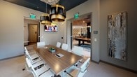 This single-floor amenity space is thoughtfully designed to blend professional functionality with upscale comfort. The centerpiece is a well-appointed conference room featuring a large wood-grain conference table surrounded by sleek white ergonomic chairs, ideal for meetings and collaborative work sessions. Overhead, striking tiered drum pendant lights in black and gold add a bold, contemporary flair, while large windows allow ample natural light and offer views of the surrounding outdoor landscape and adjacent building exteriors. A wall-mounted flat-screen TV enhances the room's utility for presentations. One accent wall is painted in a deep navy blue, complemented by a framed abstract artwork, lending the space a sophisticated and curated aesthetic. The patterned ceiling tile above the conference area adds an additional layer of visual interest. An adjoining lounge area provides a more relaxed setting, with built-in bench seating, plush accent pillows, and oval marble-top tables positioned beneath pendant glass globe lights. Large windows in this area further brighten the space. Through an open doorway, a recreation room is visible, complete with a billiards table, decorative lighting, and stylish furnishings, rounding out this versatile and well-designed communal amenity space.