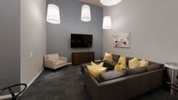 Located at 401 Granby St in downtown Norfolk, Virginia, this amenity game room offers a well-designed, multifunctional space ideal for recreation and socializing. The room features soaring ceilings that lend an open, airy feel, accentuated by a series of large white drum pendant lights suspended at varying heights throughout the space.

The game area is anchored by a full-size billiards table with a rich mahogany frame and slate playing surface, accompanied by a wall-mounted cue rack nearby. A foosball table adds additional entertainment options, while a dedicated gaming console station rounds out the interactive amenities.

A comfortable lounge area occupies one end of the room, furnished with a large sectional sofa and accent chair in neutral tones, accented with gold throw pillows. A wall-mounted flat-screen television sits above a walnut media console, creating an inviting space for relaxation. A round glass-top dining table with modern chairs provides a casual seating option.

The décor is cohesive and contemporary, with soft gray walls, dark carpeted flooring, and a distinctive grid of warm wood panel art tiles on one accent wall. Framed artwork is thoughtfully placed throughout, adding visual interest. French doors provide access and contribute to the polished, modern character of this well-appointed amenity space.