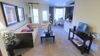 This single-floor residence showcases an open-concept layout that connects the living and dining areas in a cohesive, flowing arrangement. The living room is anchored by a tufted sofa dressed with decorative pillows, complemented by a sleek dark-wood coffee table and a coordinating console table along the wall. Plush wall-to-wall carpeting runs throughout, adding warmth and texture underfoot. Crown molding frames the ceilings, lending a refined architectural detail to the space. Multiple large windows dressed with floor-length curtains and blinds flood the interior with abundant natural light, creating an airy atmosphere. An accent wall in a deep, rich tone adds visual depth and contrast against the neutral surrounding walls. The dining area, partially visible beyond the living space, features a classic table and chair arrangement beneath track lighting, while a raised counter or bar area provides a natural transition point toward the kitchen. The overall ambiance is polished and inviting, balancing comfortable furnishings with tasteful design elements that make the space feel both functional and stylish.