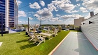 Located atop 51 S Broadway in the heart of White Plains, NY, this generously sized rooftop deck delivers an impressive open-air experience with panoramic views of the surrounding downtown skyline and tree-lined horizon. The deck is thoughtfully divided into distinct zones that cater to both relaxation and recreation. Lush artificial turf spans much of the surface, creating a vibrant, park-like atmosphere high above the city streets. Comfortable Adirondack chairs, rocking chairs, lounge sofas, and ottomans are arranged throughout to encourage both social gatherings and quiet relaxation. String lights draped overhead add a warm, inviting ambiance for evening use. A dedicated games area features a shuffleboard court and a giant Jenga set, while hammocks strung between posts offer a laid-back retreat. Raised planter boxes introduce greenery and floral accents, softening the urban setting. A sleek glass-railed perimeter provides unobstructed sightlines to the White Plains cityscape below, including prominent office towers and tree canopy stretching into the distance. Clean-lined, slate-toned architectural walls with wood-look tile flooring define a transitional service area near the rooftop access structure. The overall design balances modern urban aesthetics with approachable, resort-style comfort.