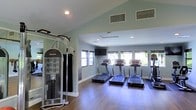 This single-floor fitness center is thoughtfully designed to provide a functional and comfortable exercise environment. The space features a row of modern treadmills positioned along a bank of windows, allowing natural light to fill the room and offering outdoor views that enhance the workout experience. A wall-mounted flat-screen television is conveniently placed for entertainment during cardio sessions. Complementing the cardio area, a multi-station cable strength training machine is positioned along the side wall, offering a variety of resistance exercises for a full-body workout. The flooring consists of durable hardwood-style planks, providing a clean and practical surface suited for a range of fitness activities. Recessed lighting in the white ceiling ensures consistent, even illumination throughout, while the soft sage-green walls contribute to a calm and focused atmosphere. HVAC vents are integrated into the ceiling for climate control comfort. The open layout and thoughtful arrangement of equipment allow for efficient use of the space, making it suitable for multiple users simultaneously. Overall, the fitness center presents a clean, modern aesthetic with all the essential amenities for an effective workout.