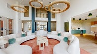 This generously sized, single-floor amenity space offers an impressive blend of contemporary design and resort-style comfort. The centerpiece of the space is a dramatic double-height lounge area anchored by bold teal and slate-gray structural columns, distinctive rope-wrapped circular chandeliers, and sweeping curved white sofas set on terracotta-hued flooring. Large windows and glass doors flood the interior with natural light while framing outdoor views, enhancing the open and airy ambiance throughout. Light wood-look tile flooring extends across much of the space, creating a seamless flow between zones. A dedicated game room features a full-size billiard table beneath a modern globe chandelier, set against soaring white walls accented with large-scale abstract artwork. Adjacent to the main lounge, a versatile dining area is appointed with a round natural wood table, woven rattan chairs, and bold green pendant lights, accompanied by counter-height bar seating along a window wall with garden views. A co-working area enclosed by glass partition walls provides functional workspace with ample natural light. Throughout, curated artwork, layered lighting, and thoughtful furnishings contribute to an elevated, design-forward atmosphere suited for both relaxation and productivity.