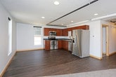 This single-floor unit combines modern finishes with an industrial aesthetic, highlighted by exposed black pipe track lighting running across textured ceilings throughout the main living areas. The open-concept living and kitchen space features dark hardwood-style flooring that creates a seamless flow between the two zones. Multiple windows along the exterior wall allow natural light to fill the room while providing outward views of the surrounding area.

The kitchen is well-appointed with warm cherry-toned flat-panel cabinetry, dark slate-style countertops, and a full suite of stainless steel appliances, including a range with over-the-range microwave, dishwasher, and refrigerator with ice maker. A double basin sink completes the functional layout.

The unit includes two carpeted bedrooms, each featuring neutral light gray walls, ceiling fans or track lighting, and ample closet space with sliding doors. The bathroom is clean and straightforward, offering a full soaking tub and shower combination with a curved rod, wood-style vinyl flooring, and a vanity with dark countertop. Throughout, light wood baseboards provide a warm contrast to the cool gray walls.