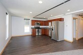 This single-floor unit combines modern finishes with an industrial aesthetic, highlighted by exposed black pipe track lighting running across textured ceilings throughout the main living areas. The open-concept living and kitchen space features dark hardwood-style flooring that creates a seamless flow between the two zones. Multiple windows along the exterior wall allow natural light to fill the room while providing outward views of the surrounding area.

The kitchen is well-appointed with warm cherry-toned flat-panel cabinetry, dark slate-style countertops, and a full suite of stainless steel appliances, including a range with over-the-range microwave, dishwasher, and refrigerator with ice maker. A double basin sink completes the functional layout.

The unit includes two carpeted bedrooms, each featuring neutral light gray walls, ceiling fans or track lighting, and ample closet space with sliding doors. The bathroom is clean and straightforward, offering a full soaking tub and shower combination with a curved rod, wood-style vinyl flooring, and a vanity with dark countertop. Throughout, light wood baseboards provide a warm contrast to the cool gray walls.
