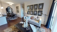 This single-floor space showcases a thoughtfully designed interior that balances sophistication with comfort. The living area serves as the centerpiece, anchored by a plush, light-toned sofa adorned with an array of decorative pillows in coordinating blues, grays, and warm neutrals. A striking gallery wall features a grid of eight framed architectural prints with dark matte backgrounds and gold frames, lending an art-forward, curated aesthetic to the space.

A tufted leather ottoman coffee table sits at the heart of the seating area, styled with candles, a decorative tray, and accent objects that add layers of visual interest. Warm hardwood flooring runs throughout, unifying the space and complementing the neutral wall tones accented by crisp white crown molding and wainscoting details.

Floor lamps with adjustable heads flank the seating arrangement, providing functional and ambient lighting. The open layout allows a clear sightline into a dining area in the background, where a round table and chairs are visible, reinforcing the connected, flowing nature of the floor plan. The overall ambiance is polished, curated, and welcoming.