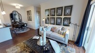 This single-floor space showcases a thoughtfully designed interior that balances sophistication with comfort. The living area serves as the centerpiece, anchored by a plush, light-toned sofa adorned with an array of decorative pillows in coordinating blues, grays, and warm neutrals. A striking gallery wall features a grid of eight framed architectural prints with dark matte backgrounds and gold frames, lending an art-forward, curated aesthetic to the space.

A tufted leather ottoman coffee table sits at the heart of the seating area, styled with candles, a decorative tray, and accent objects that add layers of visual interest. Warm hardwood flooring runs throughout, unifying the space and complementing the neutral wall tones accented by crisp white crown molding and wainscoting details.

Floor lamps with adjustable heads flank the seating arrangement, providing functional and ambient lighting. The open layout allows a clear sightline into a dining area in the background, where a round table and chairs are visible, reinforcing the connected, flowing nature of the floor plan. The overall ambiance is polished, curated, and welcoming.
