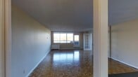 This single-floor space is characterized by its open layout and timeless design elements. The main living area is anchored by polished parquet wood flooring arranged in a classic herringbone-style pattern, adding warmth and texture underfoot. Large windows span a generous portion of the exterior wall, flooding the interior with natural light and providing pleasant views of surrounding greenery. The textured ceiling and neutral-toned walls throughout create a clean, understated backdrop well-suited to a variety of interior styles. A notable architectural detail is the elegant French door, which adds character and provides a visual transition between areas of the space. Baseboard heating units are positioned beneath the windows, ensuring efficient warmth throughout the colder months. The open floor plan allows for flexible furniture arrangements, making the living area highly adaptable. Overall, the space presents a bright and airy ambiance with classic finishes, offering a comfortable and functional environment on a single level.