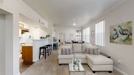This single-story, 2-bedroom, 2-bathroom home features a bright, open-concept layout that seamlessly connects the living, dining, and kitchen areas. The spacious living room is carpeted and bathed in natural light from large windows with vertical blinds, creating an airy, welcoming atmosphere accented by crown molding throughout. A peninsula counter with bar seating bridges the living area to the kitchen, which features wood-look flooring, white cabinetry with wood trim, light countertops, and a full suite of appliances in a functional galley-style configuration. The dining area sits adjacent to the living room and includes a door providing direct outdoor access, alongside a large window that brings in additional natural light. The primary bedroom is generously sized with plush carpeting, a ceiling fan, crown molding, a walk-in closet, and a private en-suite bathroom. The bathrooms feature wood-look flooring, wide vanity mirrors, light countertops, and a shower-tub combination. A second well-sized bedroom and an additional walk-in closet provide ample storage. The consistent use of neutral tones, crown molding, and plentiful natural light throughout creates a cohesive and comfortable living environment.