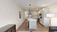 This thoughtfully designed single-floor residence offers two bedrooms and one bathroom within a compact yet efficient layout. The open-concept living, dining, and kitchen area serves as the heart of the home, featuring wood-look plank flooring that flows seamlessly throughout. The kitchen is a standout, outfitted with white shaker-style cabinetry, a subway tile backsplash, stainless steel appliances including a refrigerator, range, microwave, and dishwasher, and a central island with a single-basin sink and seating for two, illuminated by a stylish pendant light with a brass fixture.

The living area receives natural light through large windows and a glass-panel door, creating a bright and airy atmosphere. The primary bedroom continues the wood-look flooring, includes a ceiling fan, windows with exterior views, and direct outdoor access. A walk-in closet provides practical storage. The full bathroom features a white vanity with black hardware, a large mirror, and matching flooring. A dedicated laundry room houses a full-size washer and dryer, adding everyday convenience to this well-appointed residence.