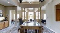 This single-story space presents an impressive lobby or reception area defined by its grand architectural character. At its center stands a substantial wooden desk with a glass top, serving as a focal reception or information point, accompanied by a freestanding chalkboard display nearby. Flanking the central axis are pairs of tall, ornate columns finished in a warm golden tone, lending a formal and distinguished aesthetic to the space.

The ceiling rises dramatically, incorporating a recessed octagonal or rotunda-style upper section adorned with lush greenery and fitted with a ceiling fan, contributing both visual interest and air circulation. Transom windows positioned high on the walls, along with large floor-to-ceiling windows and glass-paneled entry doors at the far end, allow abundant natural light to flood the interior, creating a bright and welcoming atmosphere.

Patterned carpeting in a classic floral motif extends throughout the floor, adding texture and warmth. Seating areas with chairs and tables are arranged along the perimeter, offering comfortable spaces for gathering. The overall ambiance is refined and inviting, combining traditional architectural elements with a well-appointed, communal character suited to a hospitality or community setting.