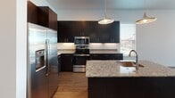This single-floor residence showcases a thoughtfully designed open-concept layout that seamlessly connects the living, dining, and kitchen areas. Rich hardwood flooring runs throughout the main living spaces, complemented by clean white walls and recessed lighting that create a bright, contemporary atmosphere. Multiple large windows flood the living area with natural light and offer views of the surrounding neighborhood, while a ceiling fan adds a touch of comfort.

The kitchen is a standout feature, finished with dark espresso cabinetry extending to the ceiling for ample storage, granite countertops, a classic white subway tile backseat, and a central island with an undermount sink. Stainless steel appliances, including a refrigerator, range, and microwave, complete the modern aesthetic. A stylish pendant light anchors the island as a focal point.

The bedroom offers a carpeted retreat with a tray ceiling detail and a dedicated ceiling fan, providing a quiet and comfortable environment. The bathroom is elegantly appointed with dark wood cabinetry, a granite-topped vanity, hardwood-style flooring, and a floor-to-ceiling marble-tiled walk-in shower enclosed by sleek sliding glass doors, delivering a spa-like experience within the home.