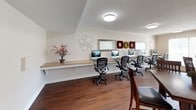 This business center offers a thoughtfully designed, single-floor workspace that balances productivity with comfort. Rich hardwood flooring flows throughout the entire space, lending warmth and a polished aesthetic to every area.

The workstation zone features a long, built-in countertop desk lined with multiple computer monitors and ergonomic mesh chairs, providing ample room for residents or guests to work efficiently side by side. Decorative wall accents, including circular metalwork and framed mirrors, add a refined touch without distracting from the functional layout.

Adjacent to the workstations is a collaborative seating area centered around a tall bistro-style table with dark wood chairs, ideal for small meetings or casual work sessions. Large windows dressed with horizontal blinds allow generous natural light to fill the room while maintaining privacy.

A comfortable lounge nook rounds out the space, furnished with a pair of patterned accent chairs accented with warm orange throw pillows, alongside a dark credenza and decorative accessories. This inviting corner provides a relaxed setting for informal conversations or a quiet break.

Flush-mount ceiling lights illuminate each zone evenly, and crown molding adds an elegant architectural finish. Overall, the business center is a versatile, well-lit, and attractively appointed space designed to meet a variety of professional and collaborative needs.