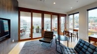 This single-floor communal space combines a fully equipped kitchen and comfortable lounge areas in a cohesive, contemporary design. The kitchen is a standout feature, showcasing floor-to-ceiling dark wood cabinetry with sleek hardware, expansive white quartz countertops, and a generous island with bar seating. Pendant lighting with glass cylinder shades hangs above the island, casting warm, focused light over the workspace. Built-in appliances and open shelving add both functionality and visual interest to the kitchen's refined aesthetic. Light wood flooring runs throughout, providing a warm contrast to the dark cabinetry.

The lounge area transitions naturally from the kitchen, furnished with leather lounge chairs, a low round coffee table, and a patterned area rug. Multiple dining tables with wood chairs are arranged near floor-to-ceiling windows, which flood the space with natural light and offer sweeping views of the surrounding urban neighborhood and distant hillside. Large glass doors open directly onto a spacious outdoor terrace, seamlessly connecting the interior with the exterior. Potted greenery and a decorative tree accent the space, adding life and warmth to the modern, sophisticated atmosphere.